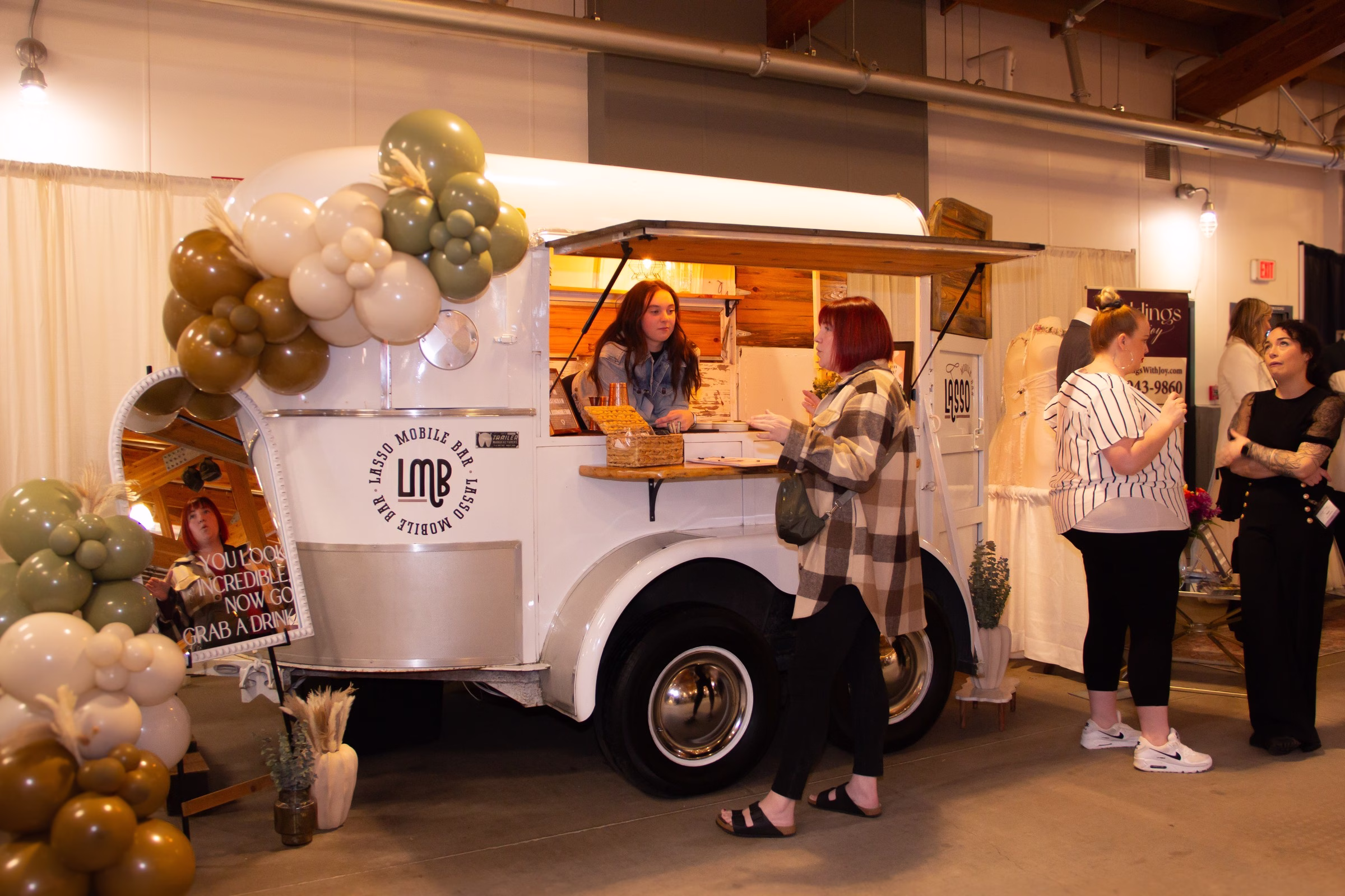 lasso mobile bar wedding vendor puyallup bridal show - Exhibiting Information Lasso Mobile Bar vintage white mobile bar trailer decorated with green and gold balloon garland serving guests at the Puyallup Wedding Show in Puyallup Washington