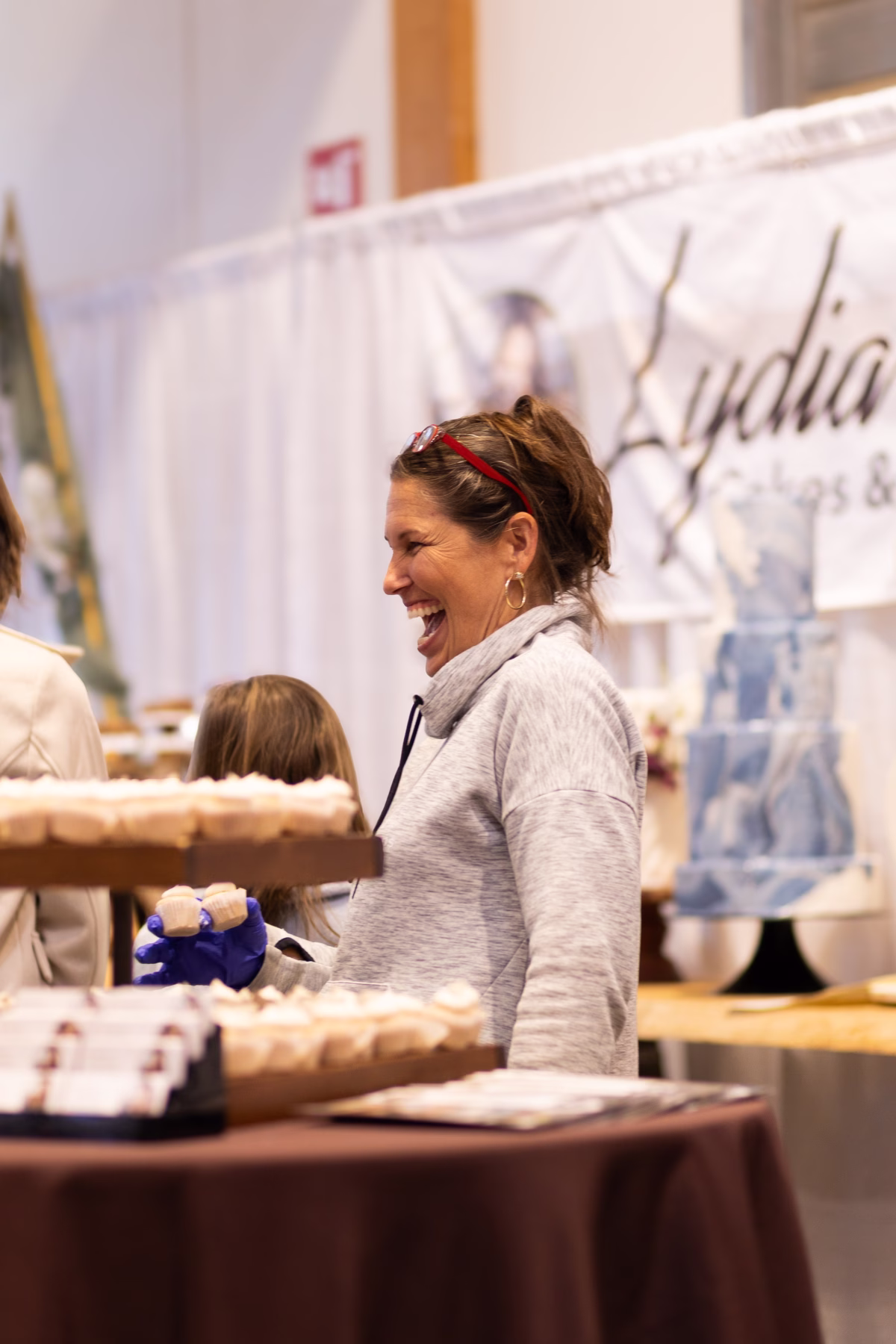 A smiling vendor in blue gloves serves cupcakes at the Lydia Cakes and Pastries booth at the Puyallup Wedding Show in Puyallup Washington