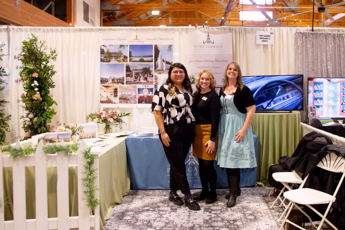 port gamble weddings events booth puyallup bridal show - Exhibiting Information Port Gamble Weddings and Events vendor booth display at the Puyallup Wedding Show in Puyallup Washington featuring three staff members posing in front of venue photos and floral arrangements