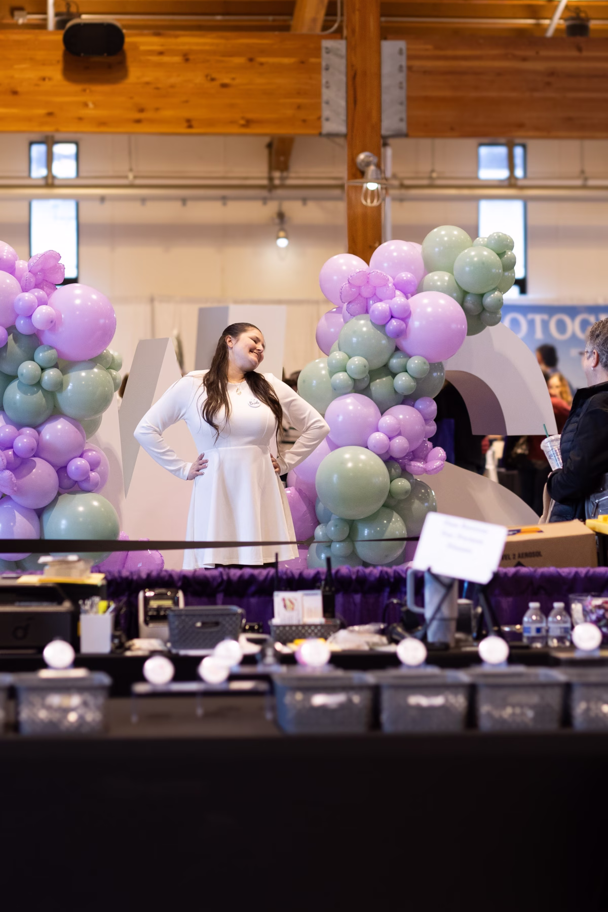 puyallup wedding show balloon arch bridal expo display - February 2027 A smiling woman in a white dress stands between a lavender and sage green organic balloon arch garland display at the Puyallup Wedding Show bridal expo in Puyallup Washington