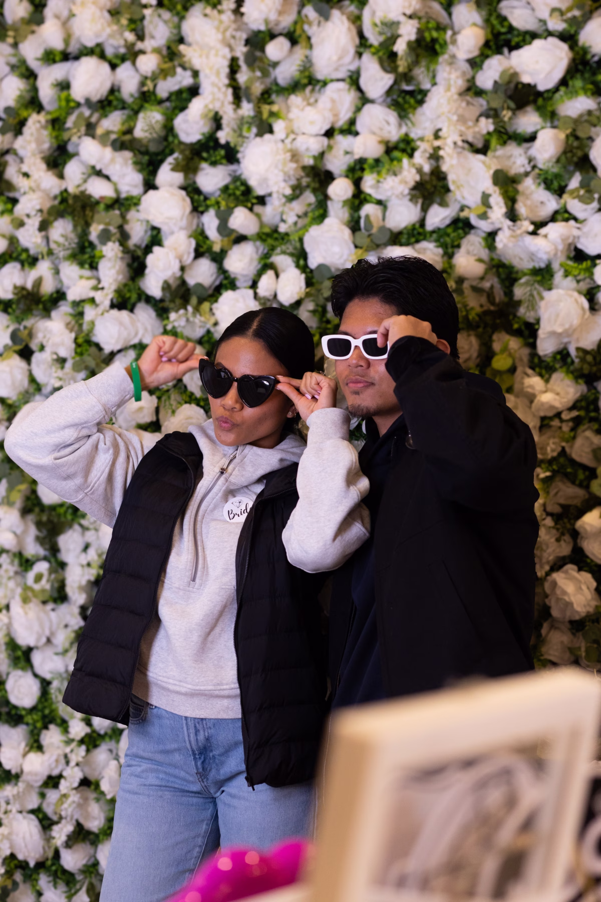 puyallup wedding show bridal couple flower wall photo - February 2027 A bride wearing a Bride button and heart-shaped sunglasses poses with a guest in front of a white floral wall backdrop at the Puyallup Wedding Show in Puyallup Washington