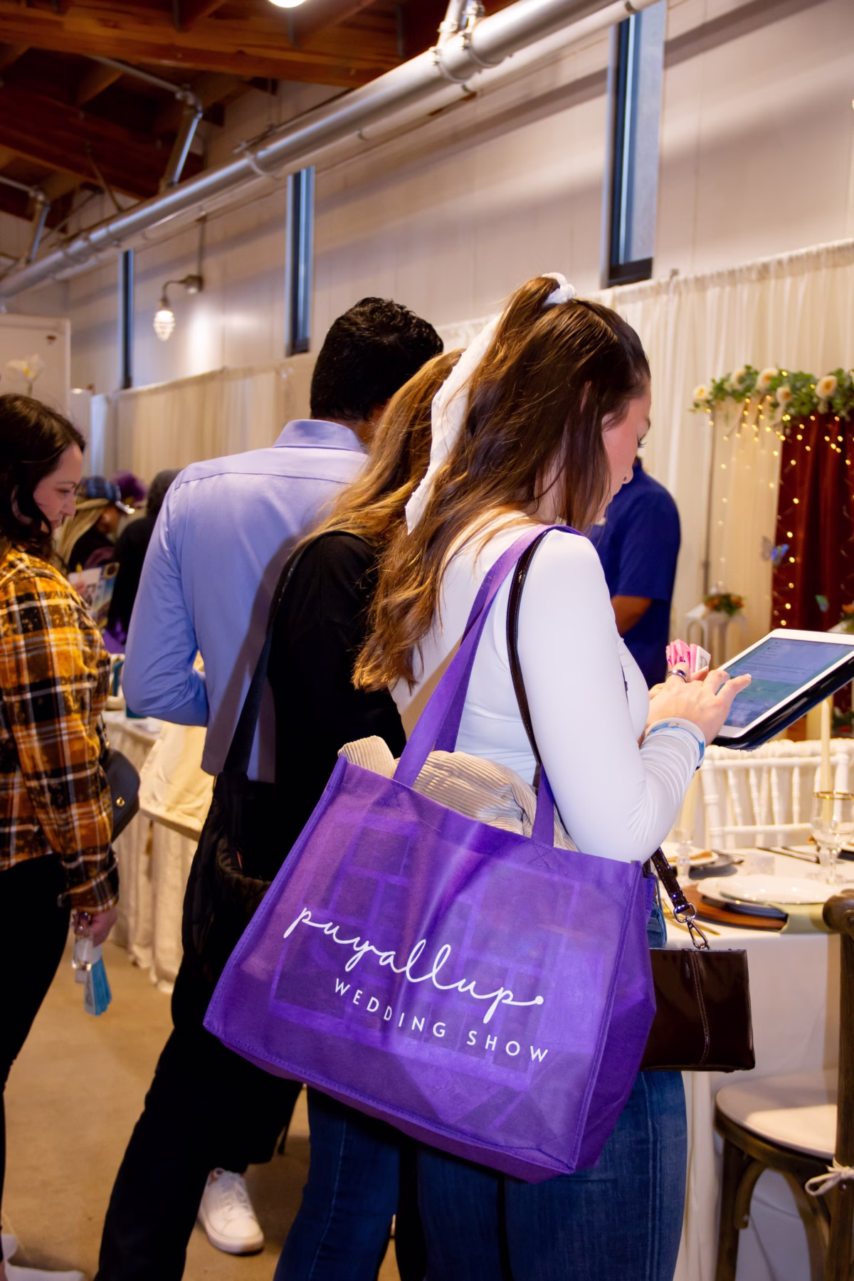 A bride attendee carrying a purple Puyallup Wedding Show tote bag browses vendor booths at the Puyallup Wedding Show bridal expo in Puyallup Washington