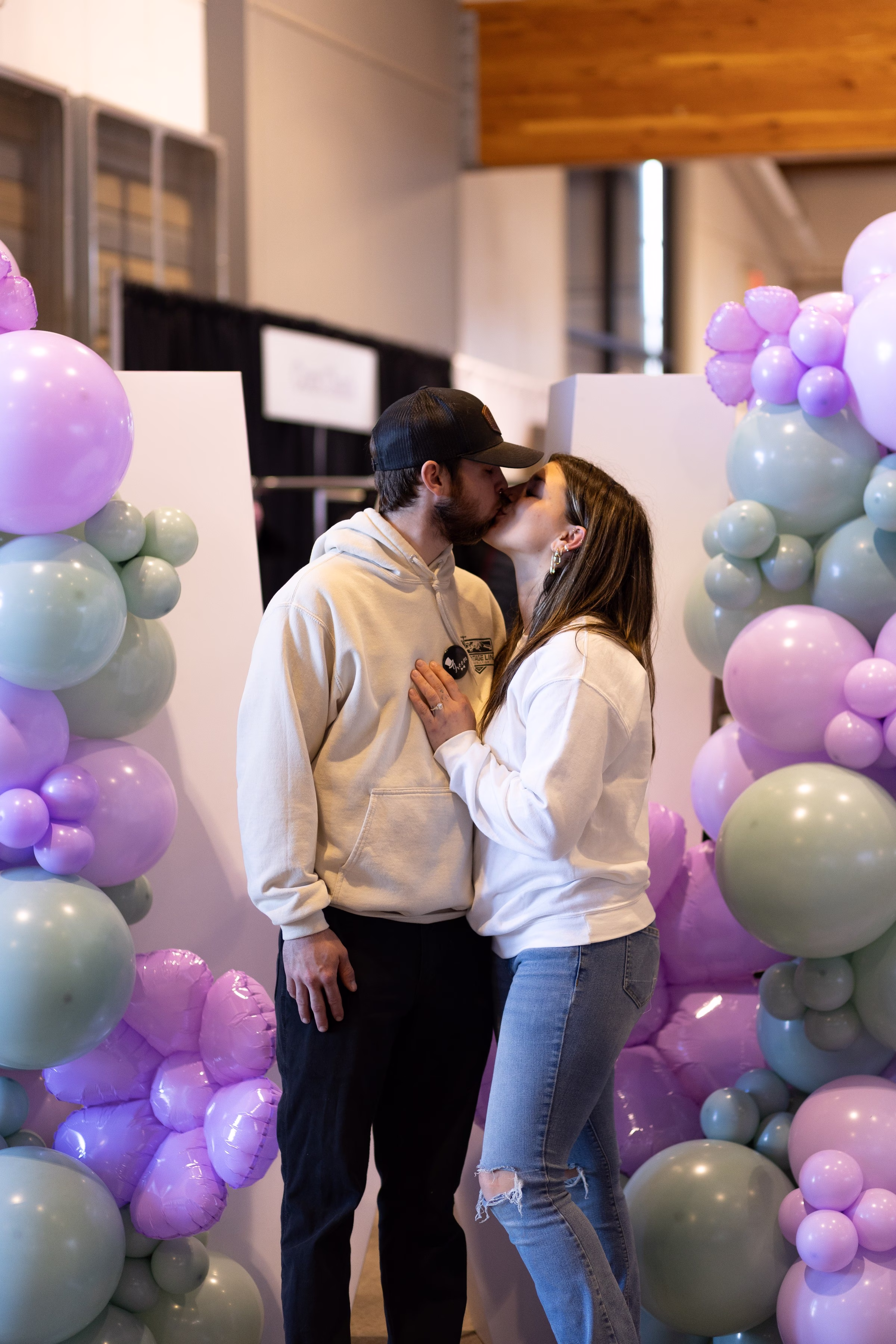 A couple shares a kiss in front of a purple and sage green balloon arch at the Puyallup Wedding Show in Puyallup Washington
